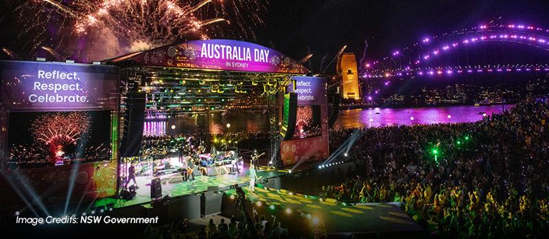 Australia Day Live concert at night with fireworks, a crowded waterfront stage, and the Sydney Harbour Bridge glowing in purple lights.