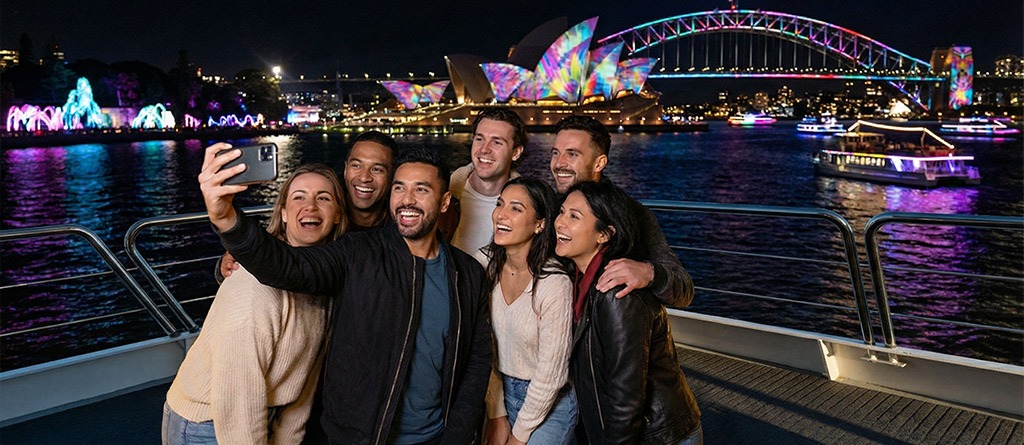 A group taking selfie from the outer decks of the Magistic Vivid Sydney Cruise, with the Vivid Light installations in the background